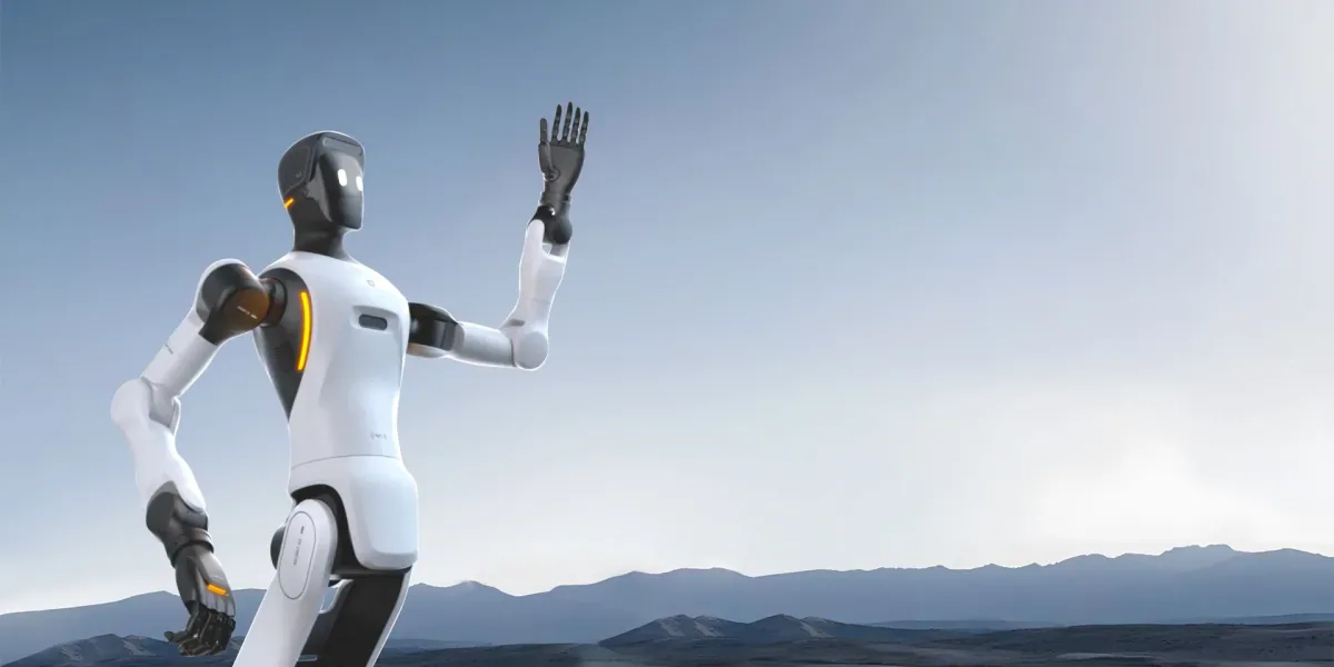 Belo-crni humanoidni robot sa narandžastim svetlosnim trakama podiže ruku u pozdrav, iza njega bledoplavo nebo i daleka brda.