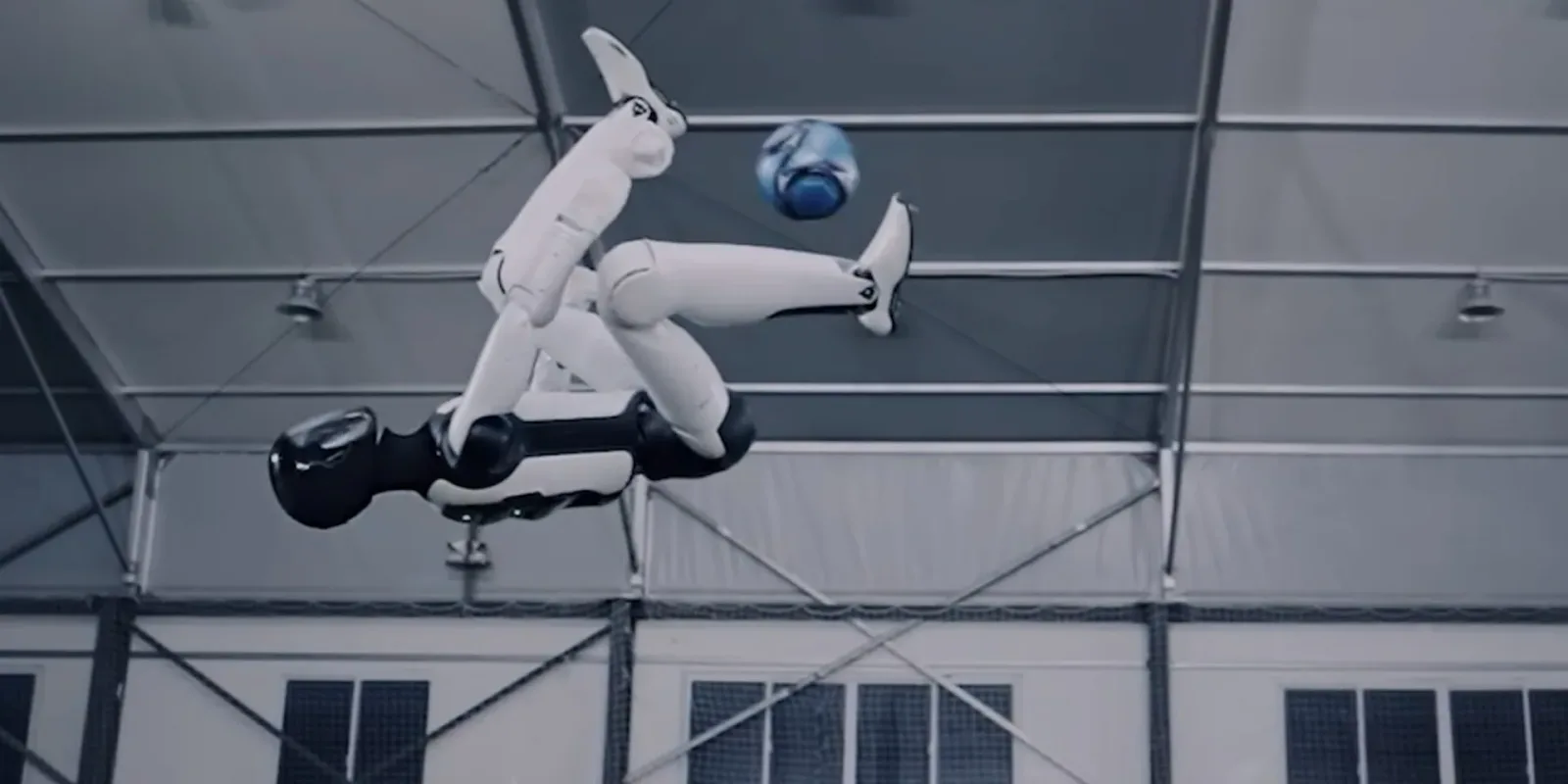 The AGIBOT A3 humanoid inverted in mid-air inside a netted training hall, one leg extended towards a ball