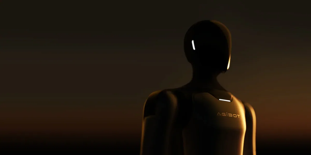 The A3 seen from the chest up against a dark horizon, its shell mostly in shadow with warm gold rim light along the head, one shoulder and the near arm, and the AGIBOT wordmark faintly lit across the chest.